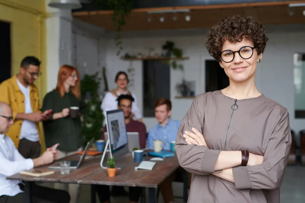 A confident businesswoman standing with arms crossed, with a team of colleagues working in the background.