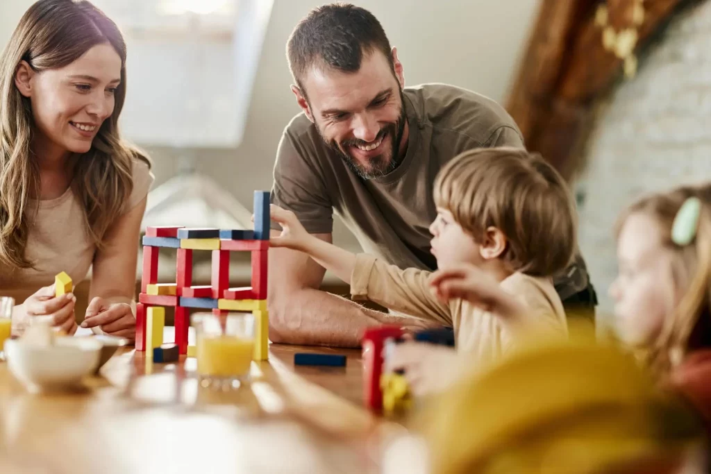 Parents playing with their two young children at a table, building with colorful blocks.