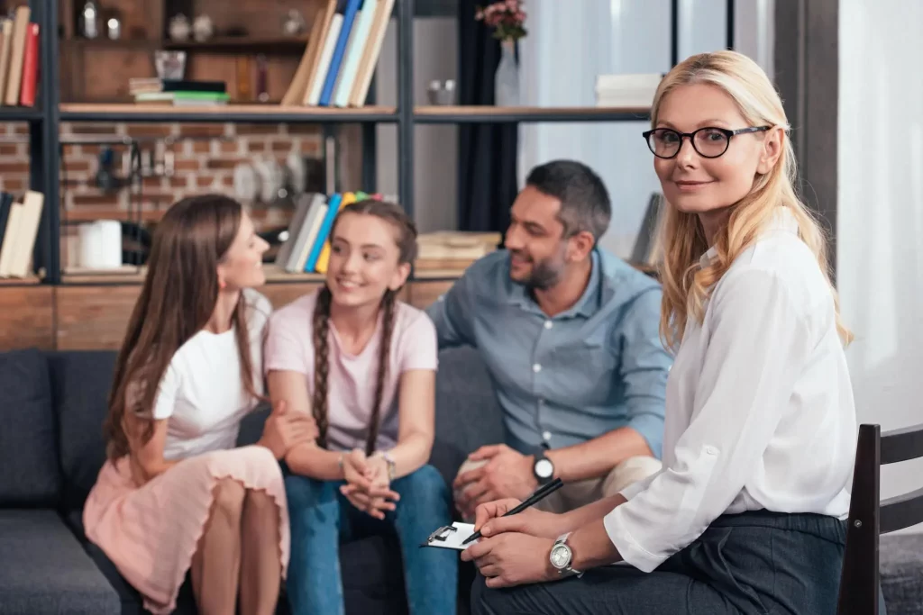 A family therapist smiling at the camera while a family of three sits on a couch in the background, talking and smiling.