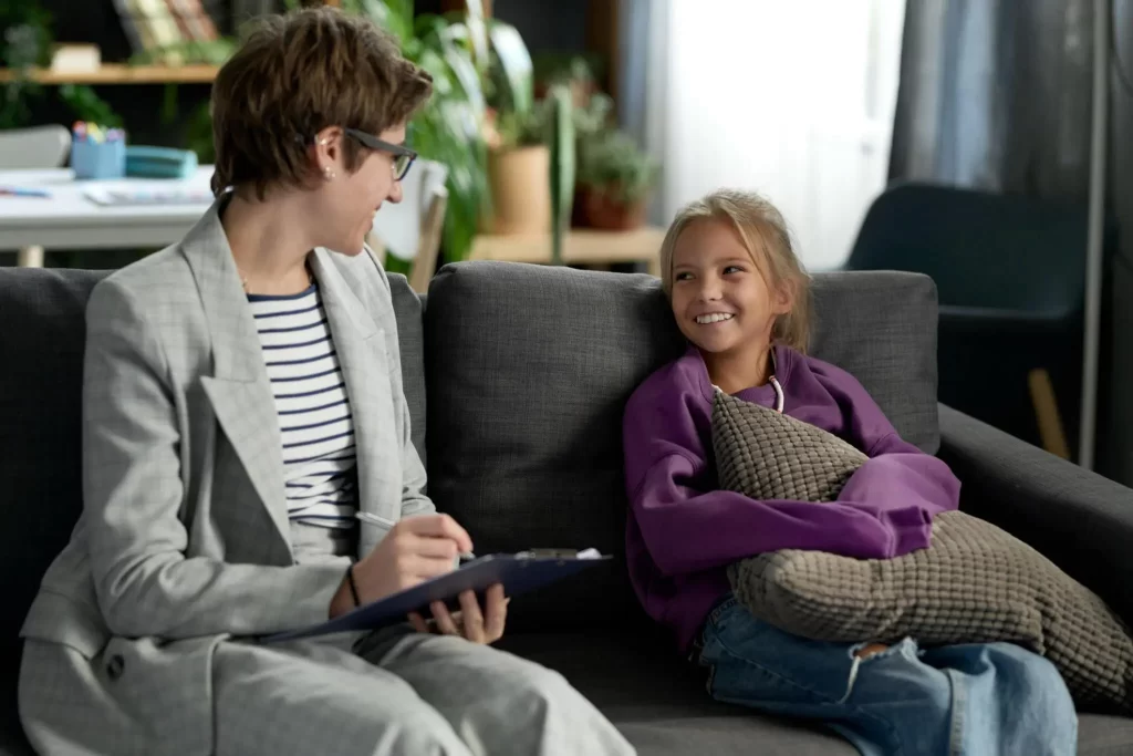 A child therapist talking with a young girl who is smiling and holding a pillow.